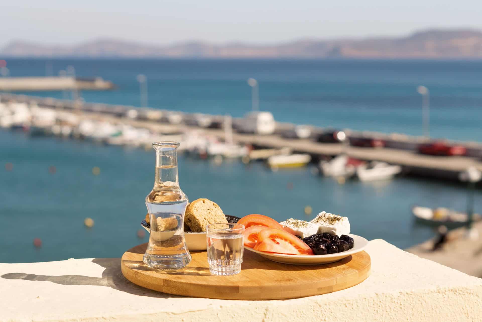Carafe de raki, pain, tomates, fromage et olives face à un port crétois